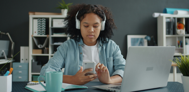Joyful office worker wearing headphones dancing in office holding smartphone working with laptop computer having fun alone in room